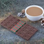 Barre de chocolat suisse au lait décorée pour Noël avec une tasse de café chaud et des pommes de pin sur une surface rustique.