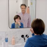 Enfant en bleu brossant ses dents dans une salle de bain moderne, parent observant derrière lui